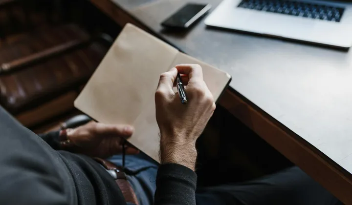 Person writing in a notebook beside a laptop at a quiet desk.